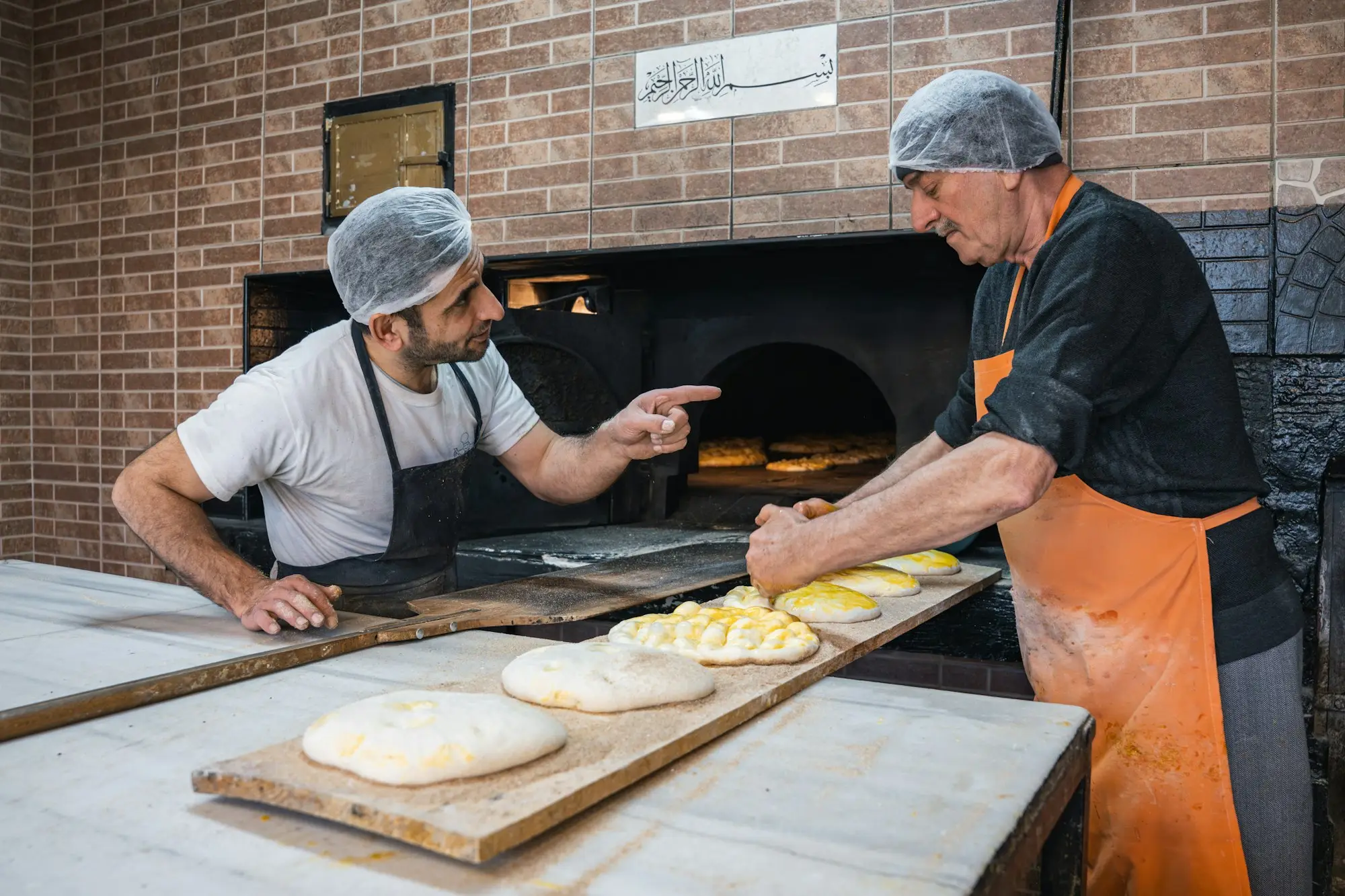 Two bakers preparing dough beside a brick oven in a busy bakery